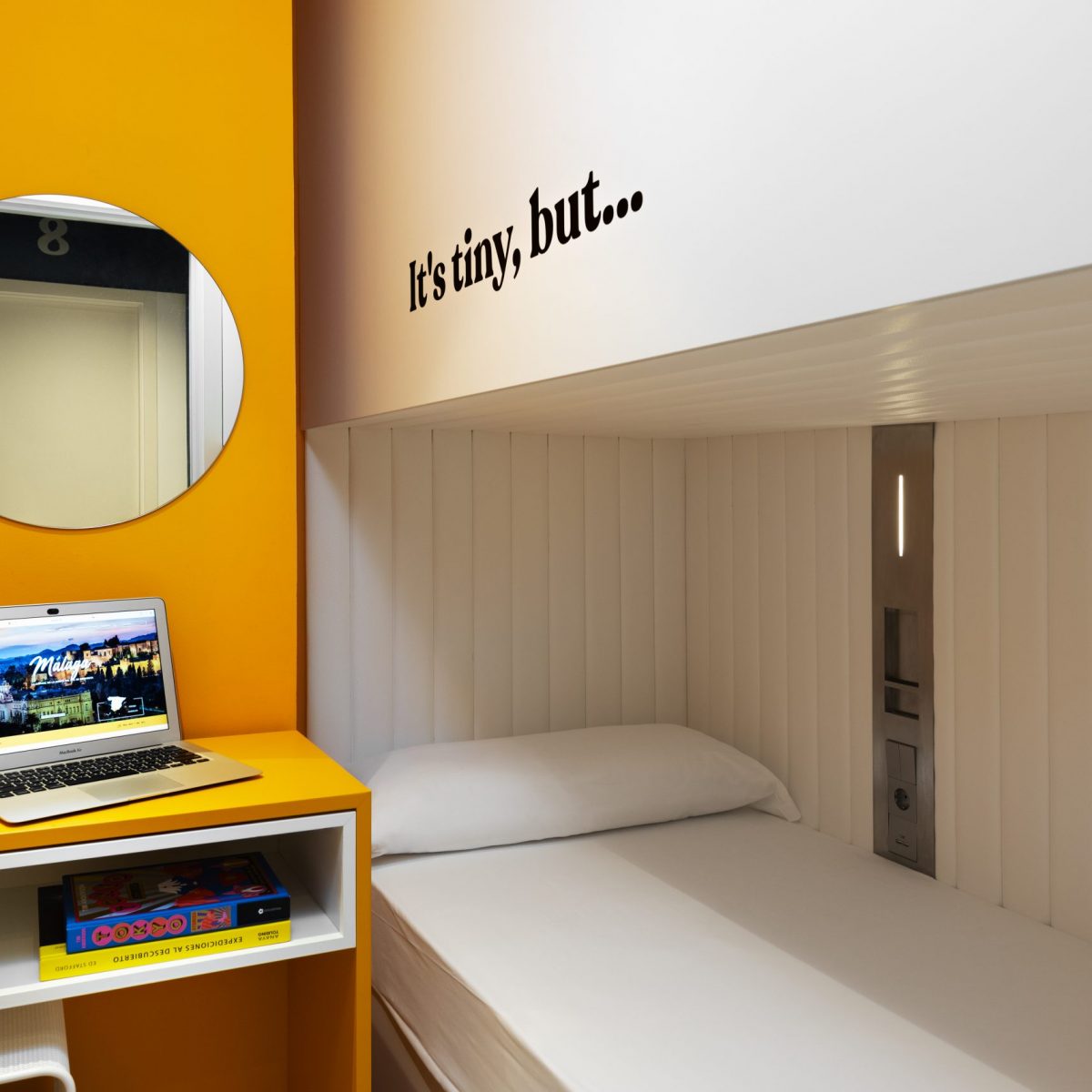 Hostel bedroom with a mirror over an orange wall and a computer on a table